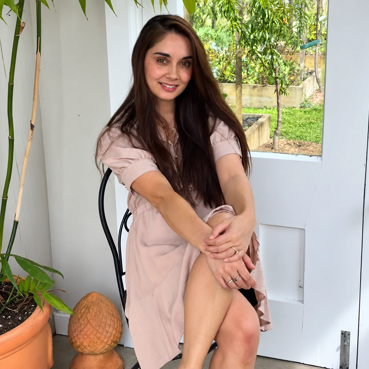 Claire seated beside plants and a window in a blush dress
